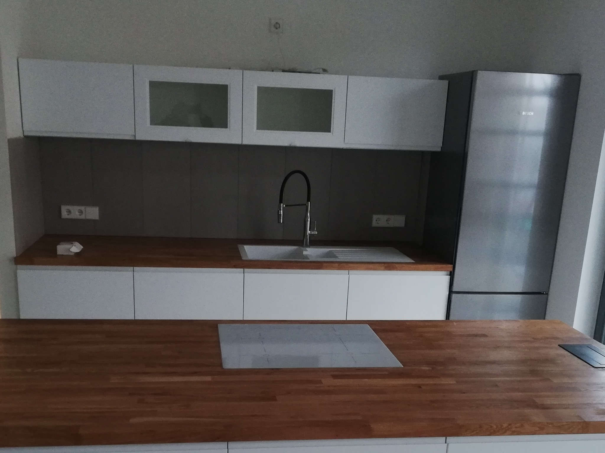 Nowoczesna kuchnia z drewnianym blatem, białymi szafkami, szarą ścianą i srebrną lodówką marki Bosch.