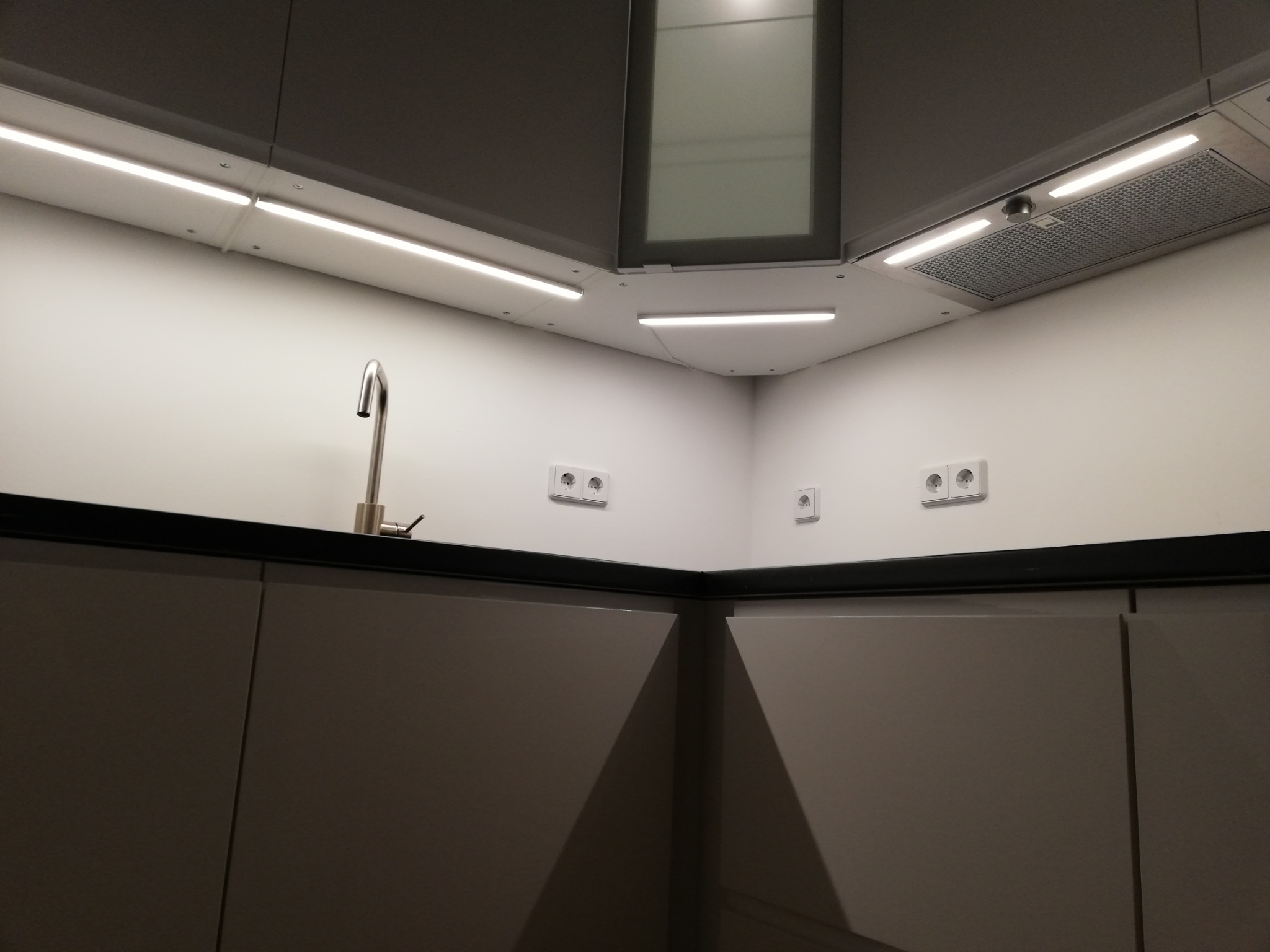 Nowoczesna, szara zabudowa kuchenna z oświetleniem LED podszafkowym, stalową baterią i gniazdkami elektrycznymi na białej ścianie.