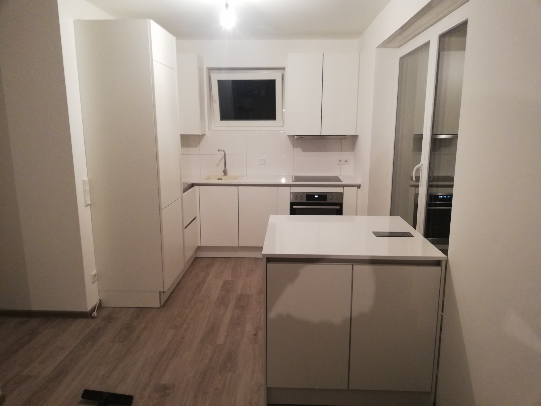 Wnętrze jasnej kuchni z białymi meblami, drewnianą podłogą i wyspą kuchenną z wbudowanym gniazdkiem elektrycznym.