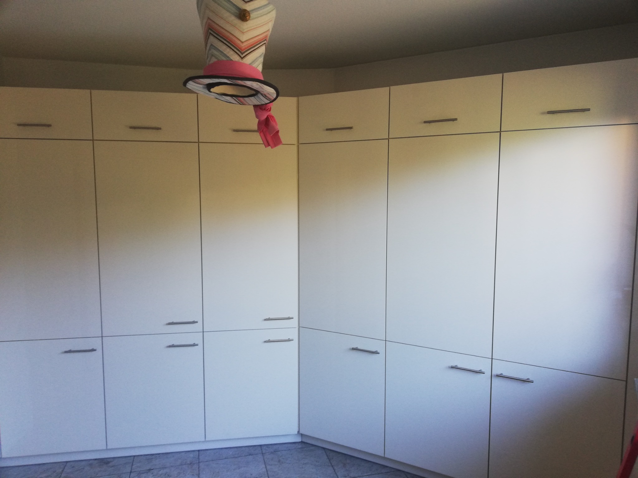 Duża, biała szafa wnękowa z metalowymi uchwytami, stojąca w rogu pokoju na szarych płytkach, z damskim manekinem głowy z kapeluszem wiszącym nad nią.