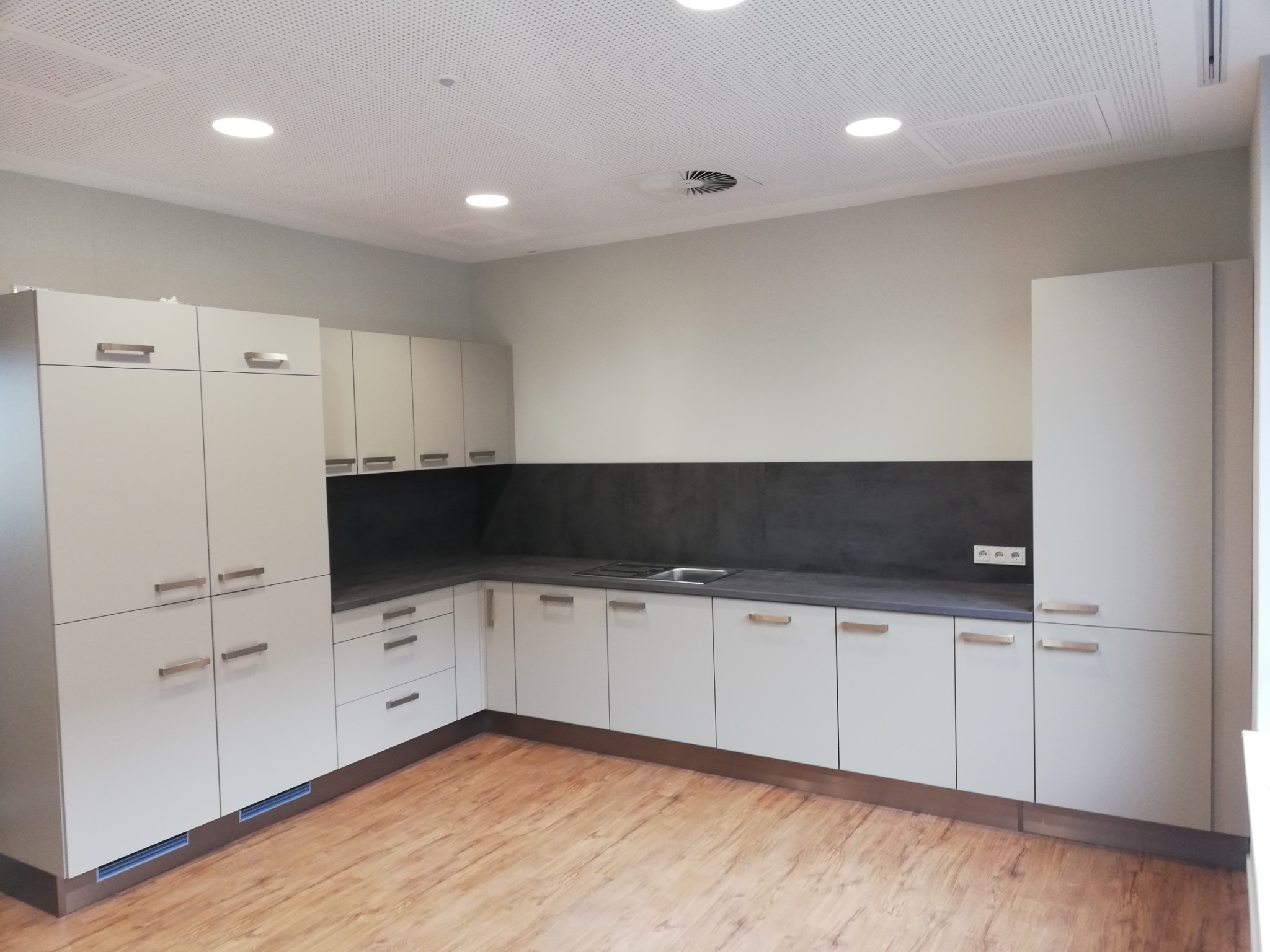 Nowoczesna, jasna kuchnia z białymi szafkami i szarym blatem, oświetlona punktowymi lampami sufitowymi, z widocznym drewnopodobnym panelem podłogowym.