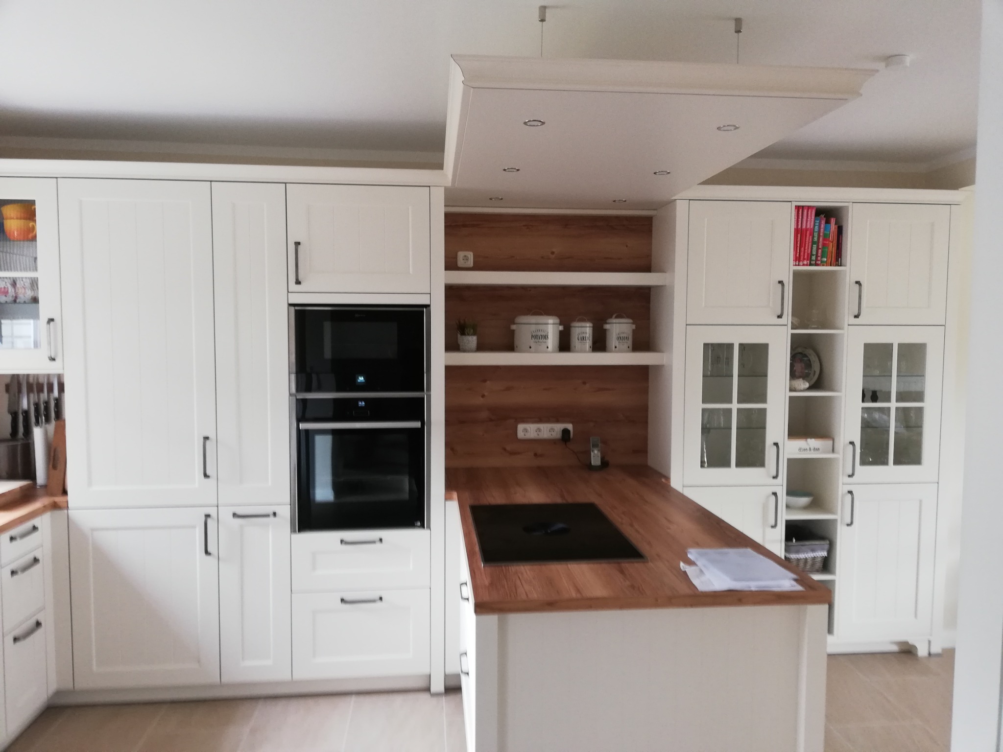 Nowoczesna biała kuchnia z drewnianym blatem i oświetleniem punktowym, widoczne szafki, piekarnik i płyta indukcyjna na wyspie kuchennej.