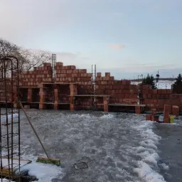 Wznoszenie ścian domu z czerwonej cegły w zimowych warunkach; widoczne zbrojenia, zapasy cegieł, oraz częściowo zamarznięta woda na fundamencie.