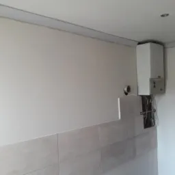 Ściana po częściowym położeniu płytek, widoczny fragment instalacji gazowej i piec, przygotowana do dalszych prac wykończeniowych.