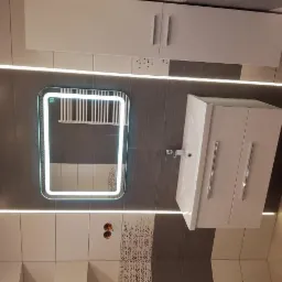 Nowoczesna łazienka z podświetlanym lustrem, białą szafką pod umywalkę i szafką wiszącą, wykończona szaro-białymi płytkami z dekoracyjnymi pasami.