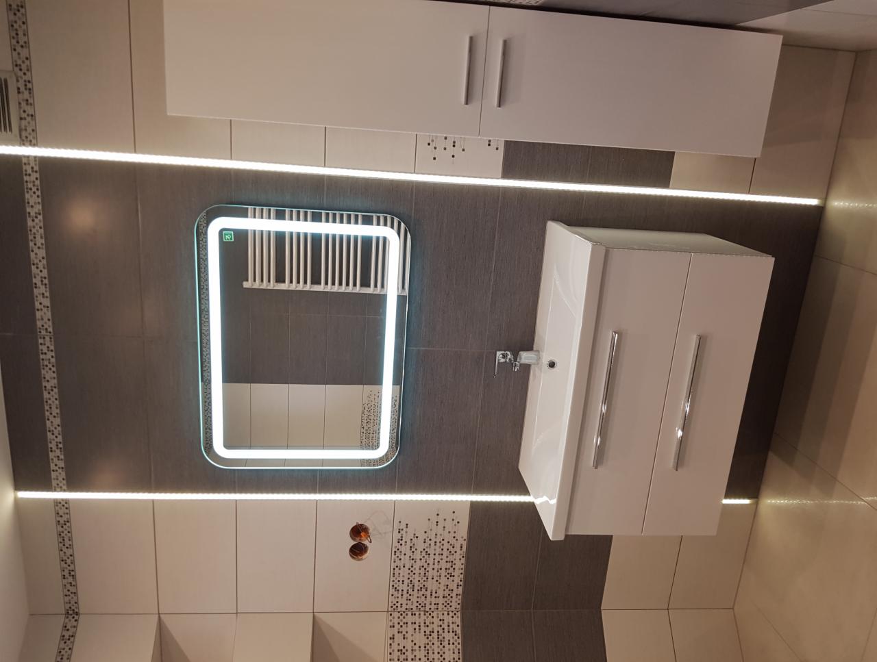 Nowoczesna łazienka z podświetlanym lustrem, białą szafką pod umywalkę i szafką wiszącą, wykończona szaro-białymi płytkami z dekoracyjnymi pasami.