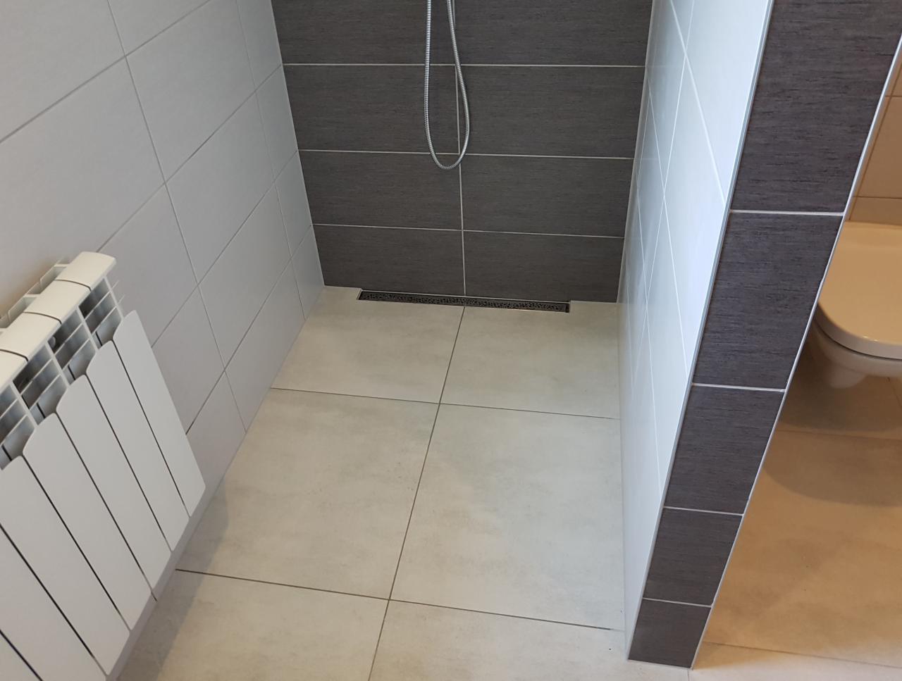 Nowoczesna łazienka z kabiną prysznicową typu walk-in, wykończona białymi i grafitowymi płytkami, z odpływem liniowym i białym grzejnikiem żeberkowym.