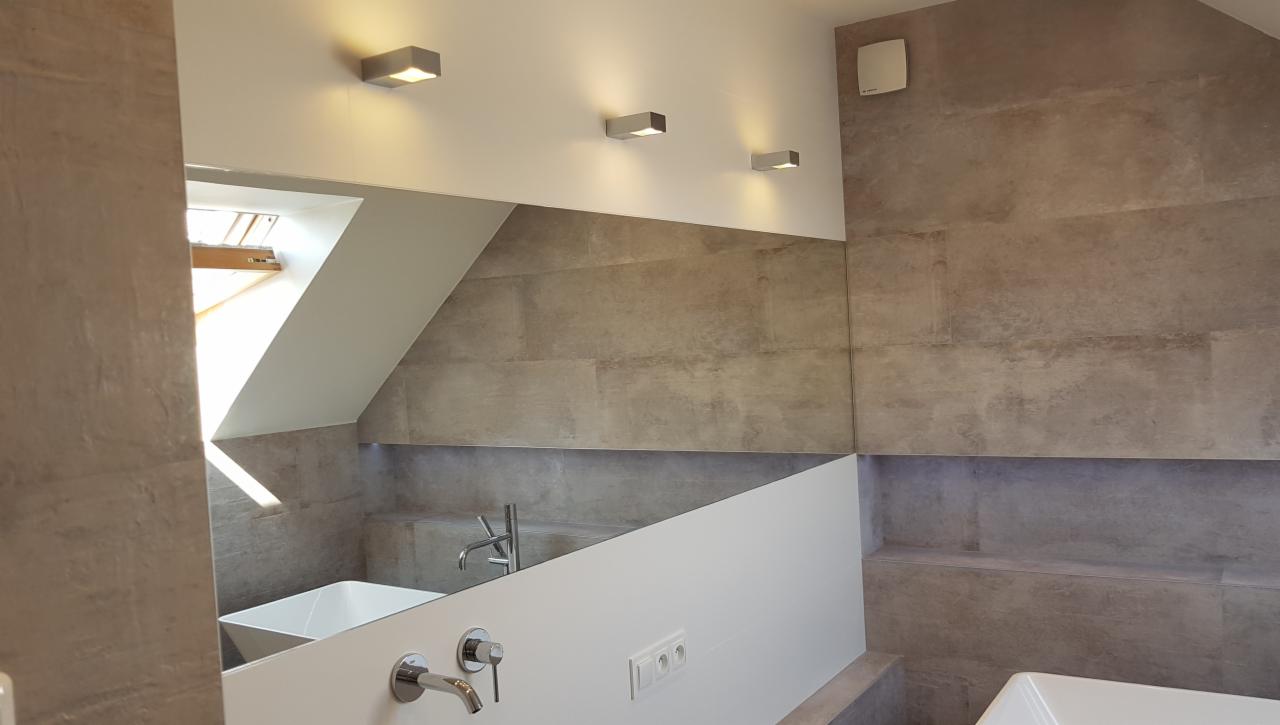 Nowoczesna łazienka na poddaszu z szarymi płytkami, oświetleniem punktowym i widocznym fragmentem wanny oraz armatury, odbicie w lustrze.