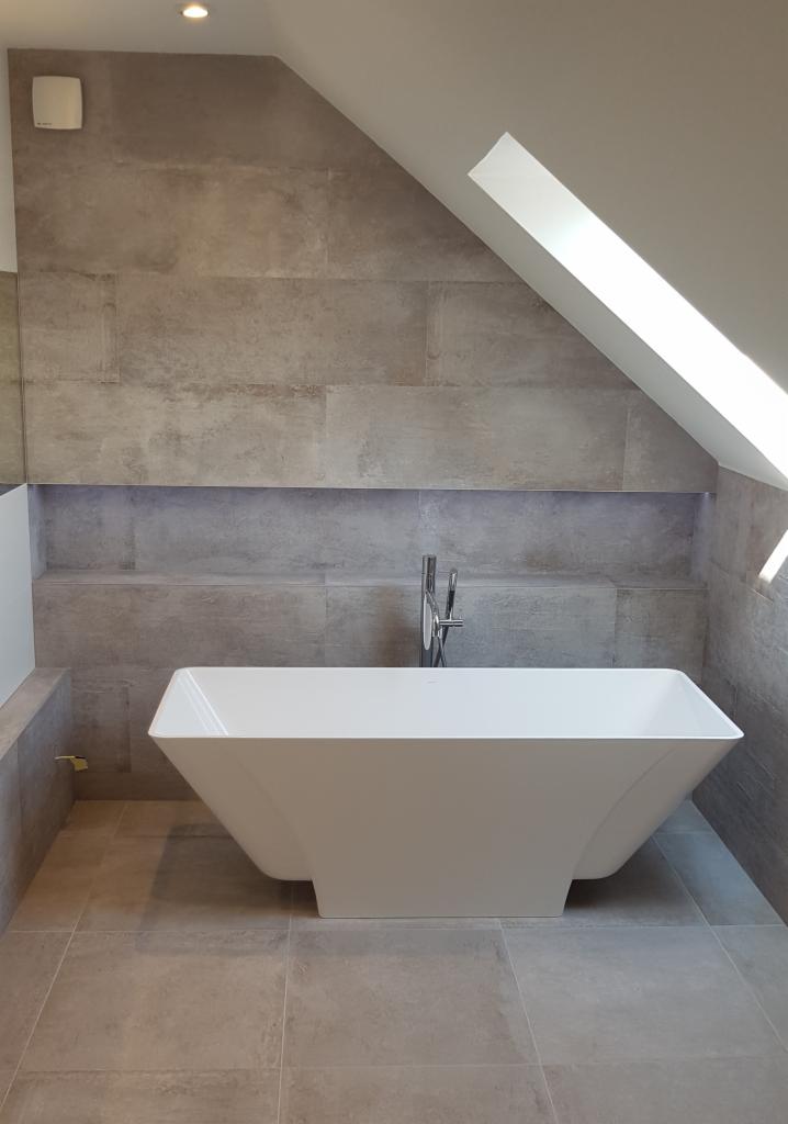 Nowoczesna łazienka na poddaszu z wolnostojącą białą wanną o geometrycznym kształcie, szare płytki imitujące kamień na ścianach i podłodze, oraz wnęka z podświetleniem LED.