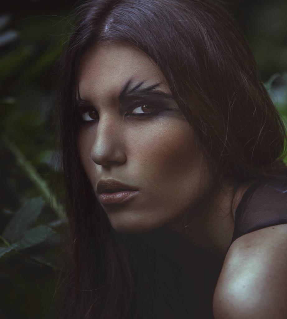 Portret kobiety z mocnym, graficznym makijażem oczu w kształcie piór, na tle rozmytej zieleni liści.