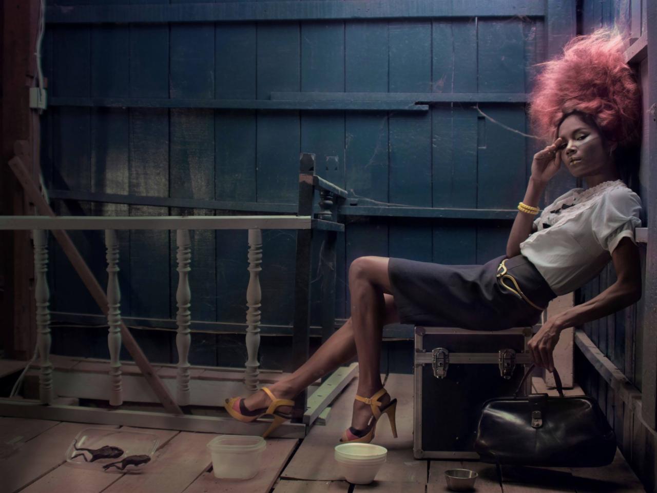 Kobieta z burzą rudych włosów pozuje siedząc na kufrze w ciemnym pomieszczeniu, obok niej leżą żaby w plastikowym pojemniku, na sobie ma elegancką bluzkę i spódnicę.