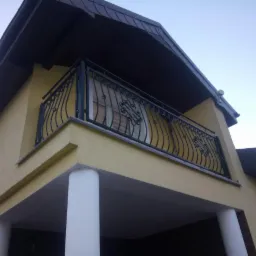 Żółty dom z balkonem zabezpieczonym czarną, metalową balustradą o dekoracyjnych wzorach, widok z dołu.