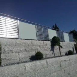 Nowoczesna balustrada z elementami szklanymi i metalowymi zamontowana na murze oporowym z granitu, z widocznymi nasadzeniami roślinnymi w tle.