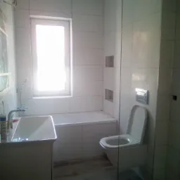 Jasna łazienka z białymi płytkami, umywalką na szafce, wanną obudowaną płytkami, toaletą podwieszaną i szklanym panelem prysznicowym. Widoczne detale wykończeniowe w postaci wnęk w ścianie.