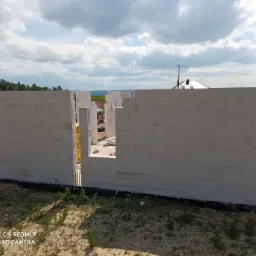 Wznoszone ściany parteru domu z białych bloczków na fundamencie z widoczną izolacją przeciwwilgociową, w tle zielony krajobraz i błękitne niebo.