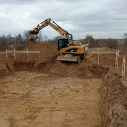 Żółta koparka CAT M313D wybiera ziemię z wykopu pod fundamenty, widoczne drewniane szalunki, w tle pole i pochmurne niebo.