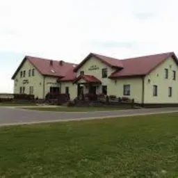 Dwukondygnacyjny budynek z ocieplonym poddaszem, pomalowany na jasnożółty kolor z czerwonym dachem, widoczny z perspektywy trawnika przed budynkiem, w tle pole uprawne.