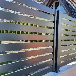 Nowoczesne ogrodzenie poziome z szarych, metalowych paneli na betonowej podmurówce, widok z zewnątrz posesji, w tle zielone tuje i dom z brązową dachówką.