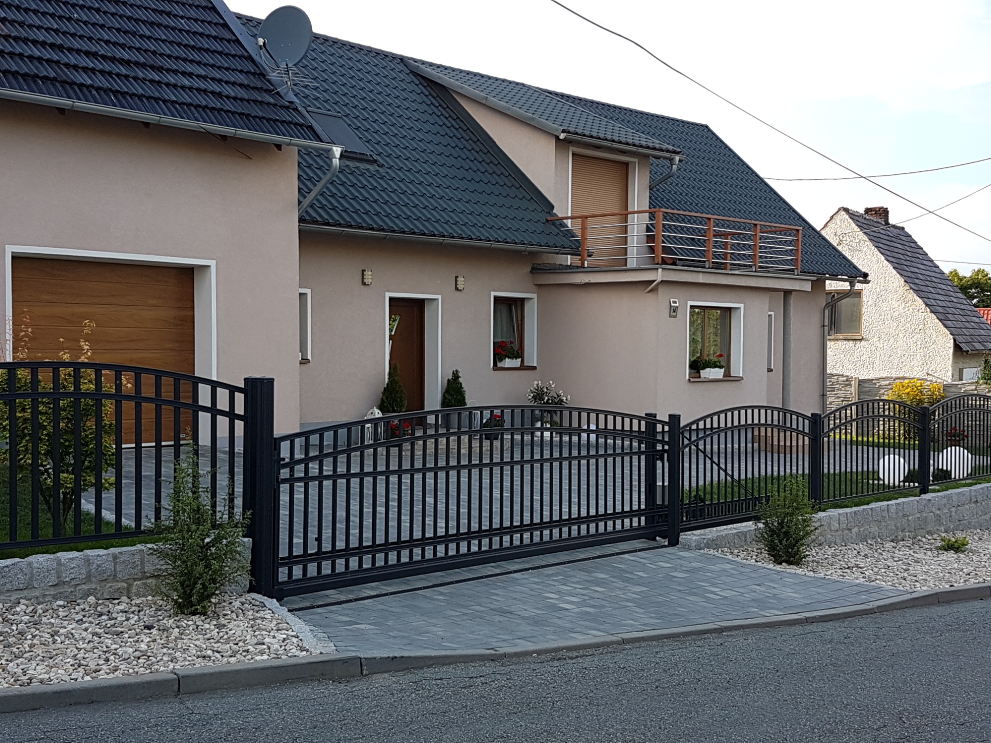 Czarna, przesuwna brama wjazdowa z pionowymi szczebelkami i łukowym wykończeniem, zamontowana przed nowoczesnym domem z szarą kostką brukową i kamiennym murkiem oporowym.