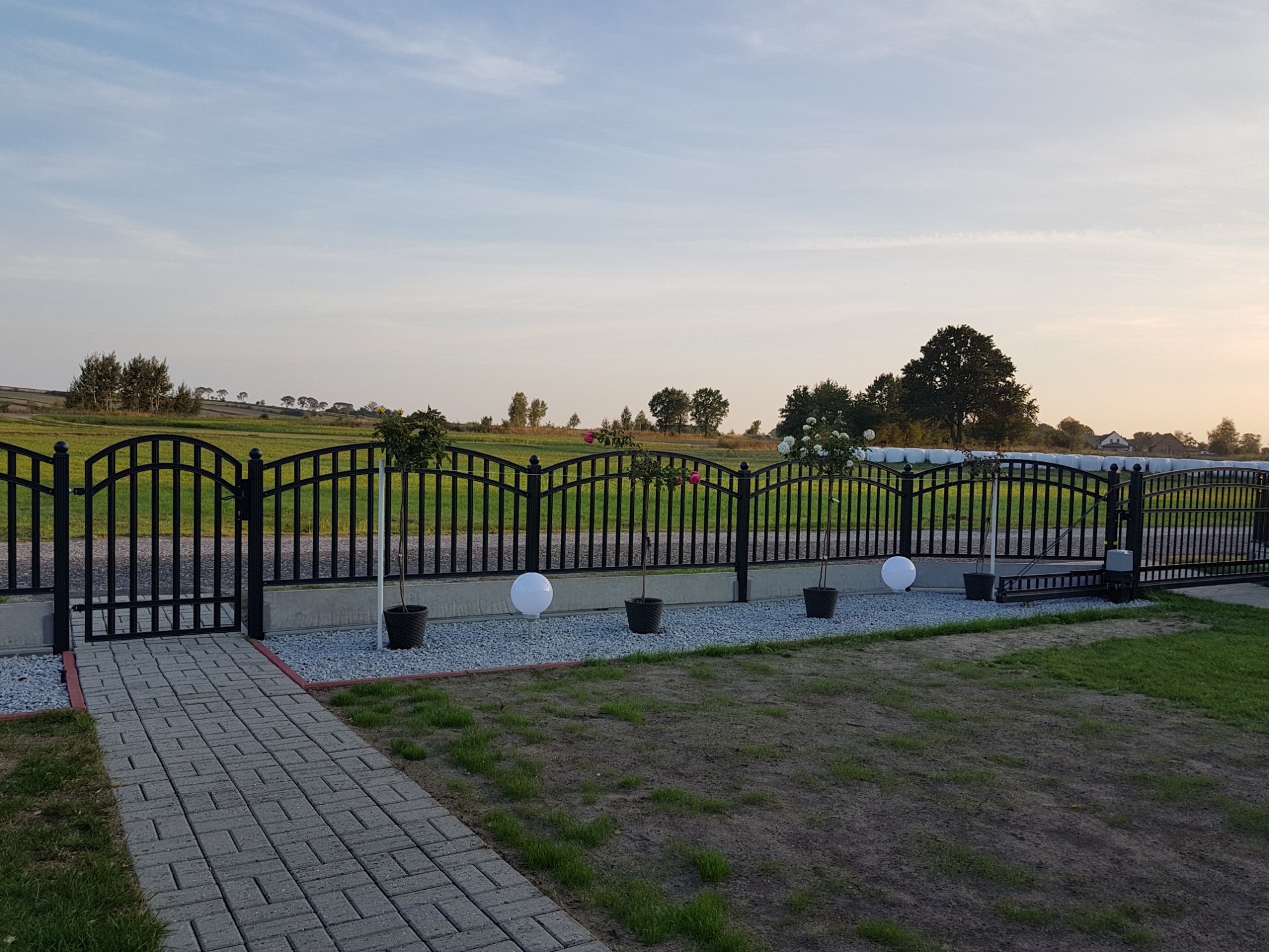 Czarny, łukowy system ogradzający z furtką i bramą przesuwną, uzupełniony o kuliste lampy ogrodowe i donice z roślinami, na tle rozległego krajobrazu.