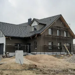 Dwukondygnacyjny dom w trakcie budowy z szarą elewacją i czarną dachówką, częściowo ocieplony białym styropianem, otoczony rusztowaniami, z widocznymi materiałami budowlanymi na działce.