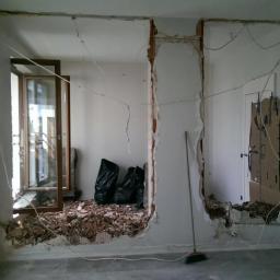 Renovation Group - Demontaż ściany działowej w mieszkaniu: widoczne uszkodzenia ścian, gruz, worki na śmieci i częściowo otwarte okno balkonowe.