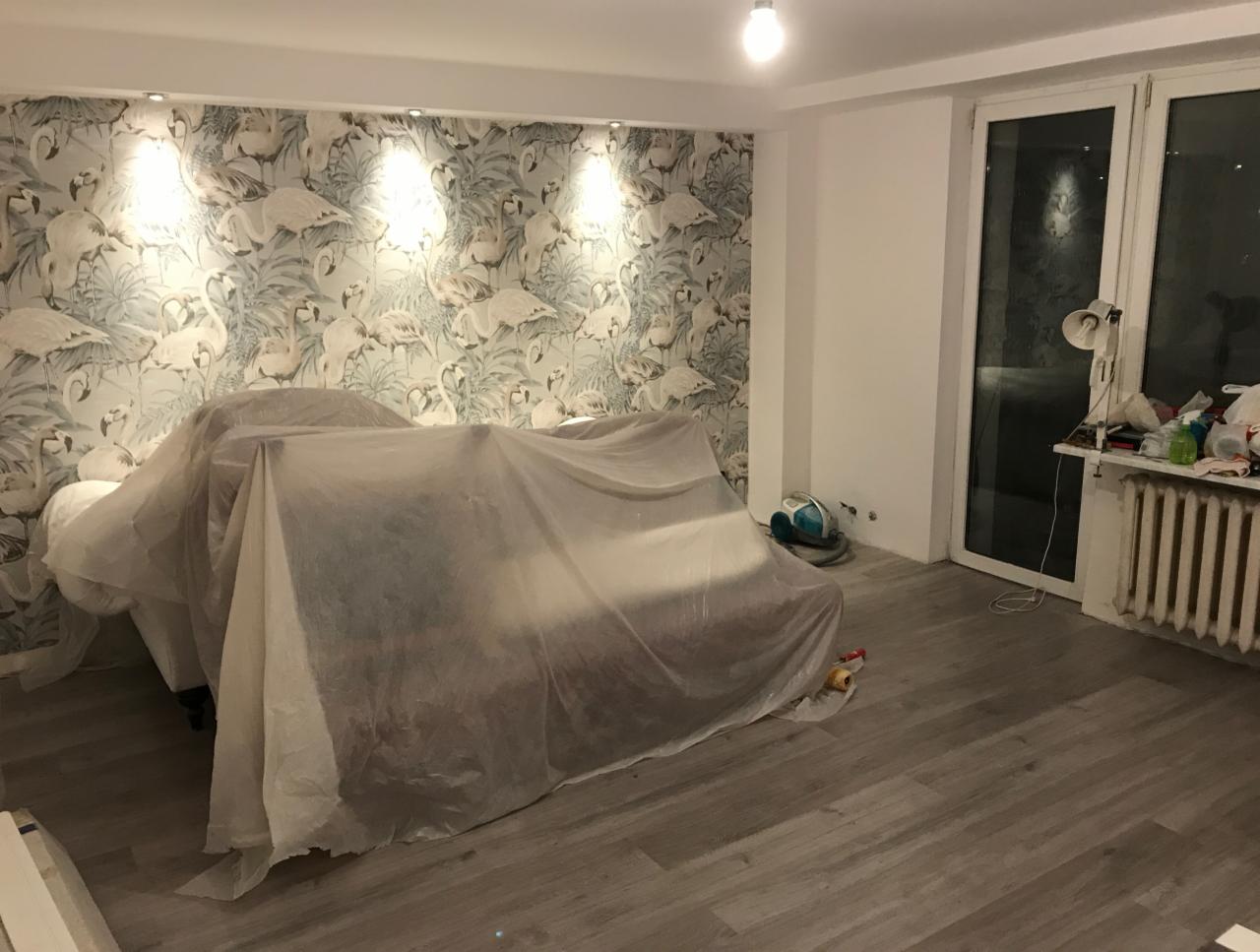 Pokój w trakcie prac: meble przykryte folią ochronną, świeżo położone panele podłogowe, tapeta z motywem flamingów na ścianie, widoczny grzejnik i akcesoria malarskie przy oknie.