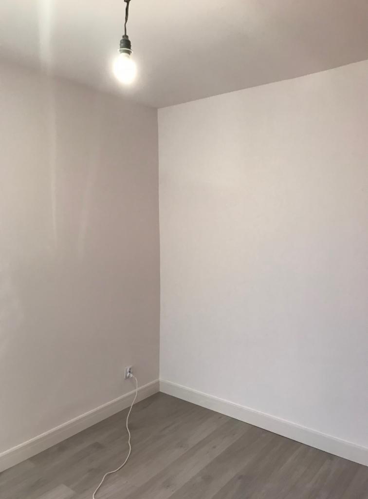 Pomalowane na biało ściany w rogu pokoju z szarą podłogą imitującą drewno, widoczna żarówka na kablu i listwy przypodłogowe.