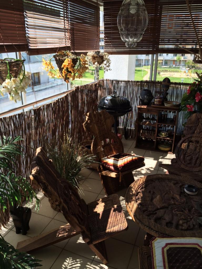 Zadaszony balkon z bambusową osłoną, drewnianymi meblami z rzeźbieniami, wiszącymi kwiatami w koszach, grillem i lampą w stylu vintage.