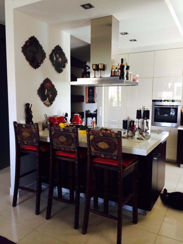 Nowoczesna, jasna kuchnia z białymi szafkami i wyspą z białym blatem, otoczona trzema wysokimi krzesłami barowymi z ciemnego drewna z czerwonymi siedziskami i zdobieniami. Na ścianie wiszą trzy...