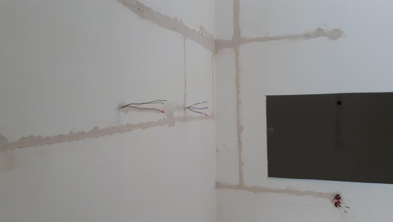 Biała ściana w trakcie bruzdowania pod instalację elektryczną, widoczne kable wystające ze ściany i wycięty otwór na puszkę, ślady gipsu wzdłuż wyżłobionych kanałów.
