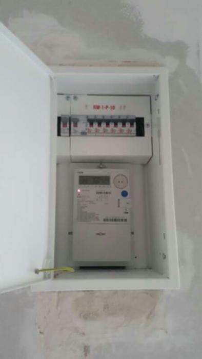 Otwarta skrzynka elektryczna z zamontowanym licznikiem energii i zabezpieczeniami, widoczna ściana w trakcie remontu.