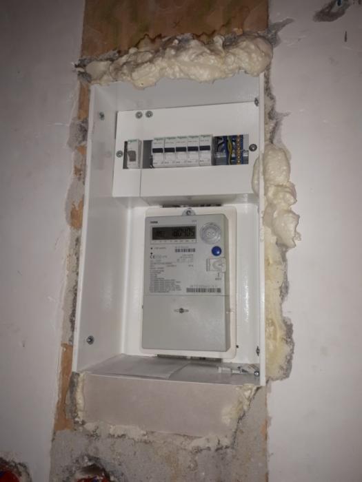 Wnęka z szarą ścianą, w której zamontowano białą skrzynkę z licznikiem elektrycznym i bezpiecznikami, szczeliny wypełnione pianką montażową.