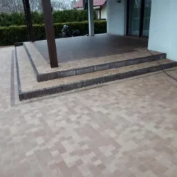 Ułożona kostka brukowa w odcieniach beżu i brązu tworząca taras z dwoma stopniami, widoczne osoby pracujące w tle, drewniana konstrukcja zadaszenia.