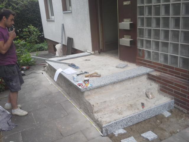 Montaż granitowych schodów wejściowych do domu, widoczny glazurnik palący papierosa, narzędzia i materiały budowlane na powierzchni schodów, w tle ściana budynku z oknami i luksferami.