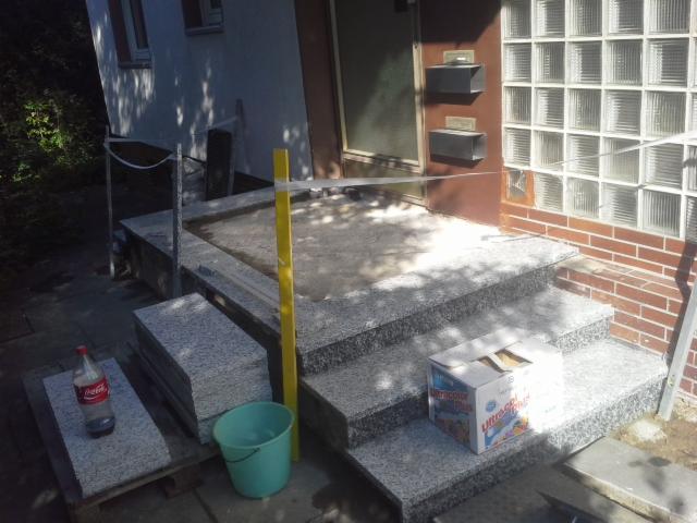 Montaż granitowych schodów zewnętrznych przed wejściem do budynku, widoczny poziomica, puste wiadro i napój w butelce na jednym ze stopni.