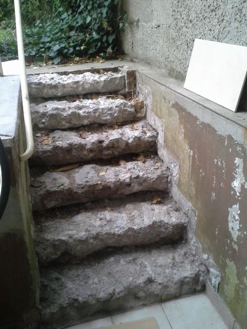 Schody zewnętrzne z surowego kamienia w trakcie remontu, z widocznym fragmentem białej płytki i śladami starej farby na ścianie.