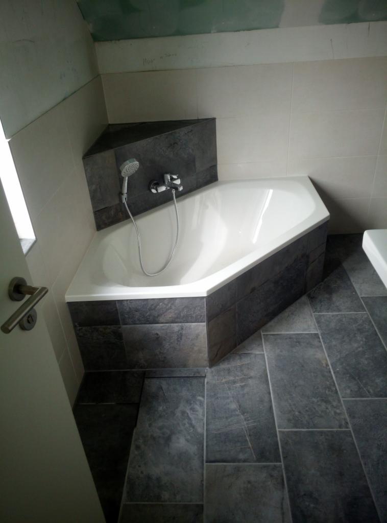 Narożna wanna obudowana ciemnymi, grafitowymi płytkami, dopasowane płytki podłogowe, bateria wannowa z prysznicem, widoczny fragment niedokończonej ściany z zielonymi płytami gipsowo-kartonowymi.