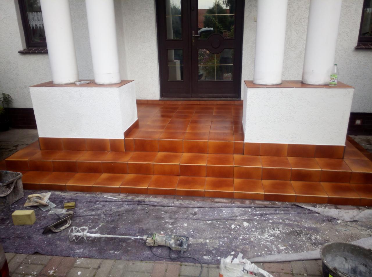 Brązowe płytki na schodach wejściowych do domu, widoczne kolumny, narzędzia glazurnicze i ślady po pracy.