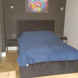 Sypialnia z łóżkiem z szarym wezgłowiem i niebieską narzutą, nad nim kolorowy portret kobiety, parkiet w jodełkę i ciemnoszare ściany.