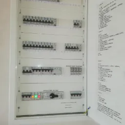 Tablica rozdzielcza z opisami obwodów, widoczne wyłączniki nadprądowe, ogranicznik przepięć, wyłącznik główny oraz listwy z opisami obwodów i zaleceniami dotyczącymi przeglądu instalacji.