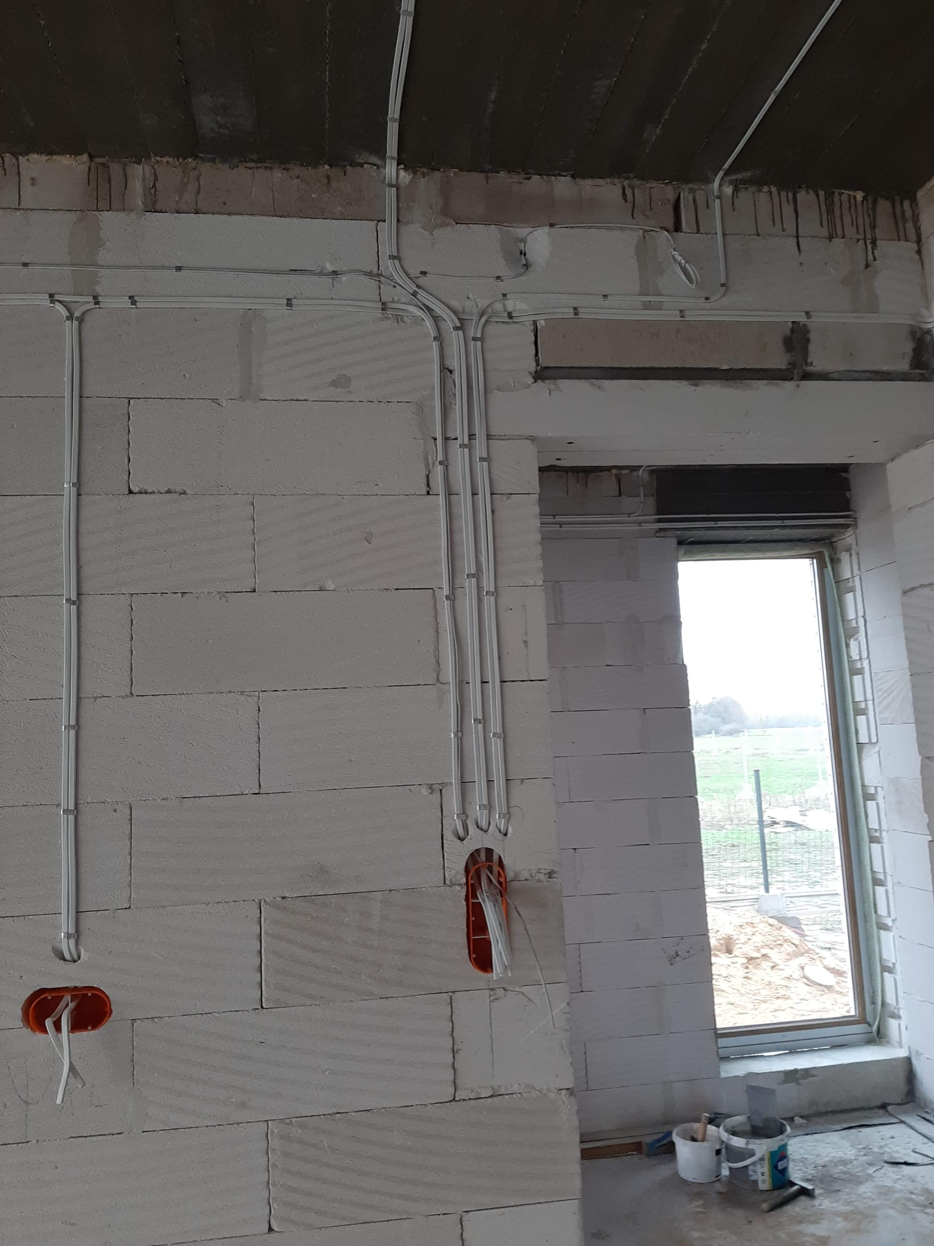 Surowa ściana z betonu komórkowego z widocznymi, szarymi rurkami instalacji elektrycznej, prowadzonymi natynkowo, z puszkami instalacyjnymi i wystającymi przewodami, okno z widokiem na zewnątrz.