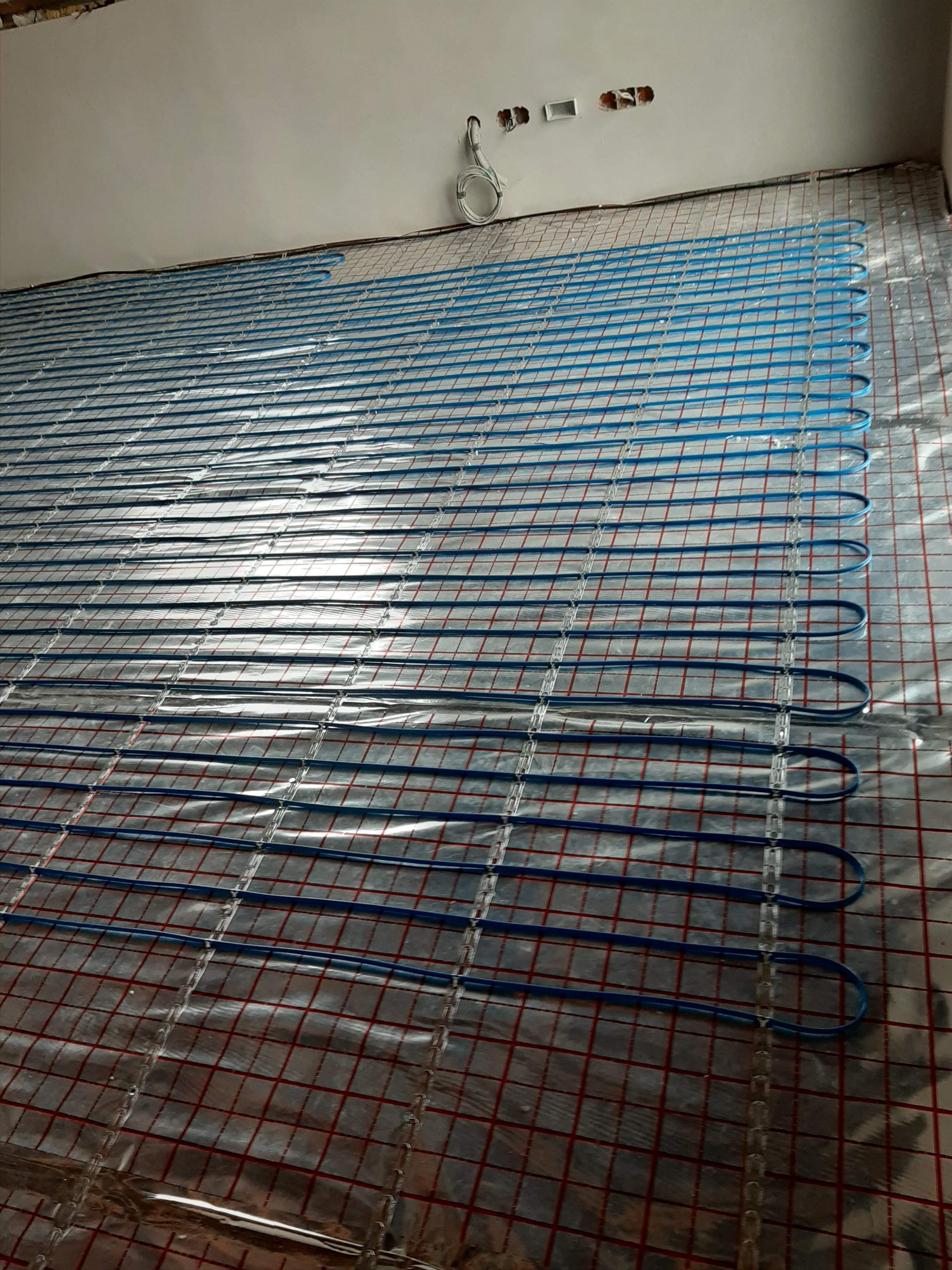 Rozłożona instalacja ogrzewania podłogowego z niebieskich rurek na folii aluminiowej z czerwoną siatką, widoczne kable wychodzące ze ściany.