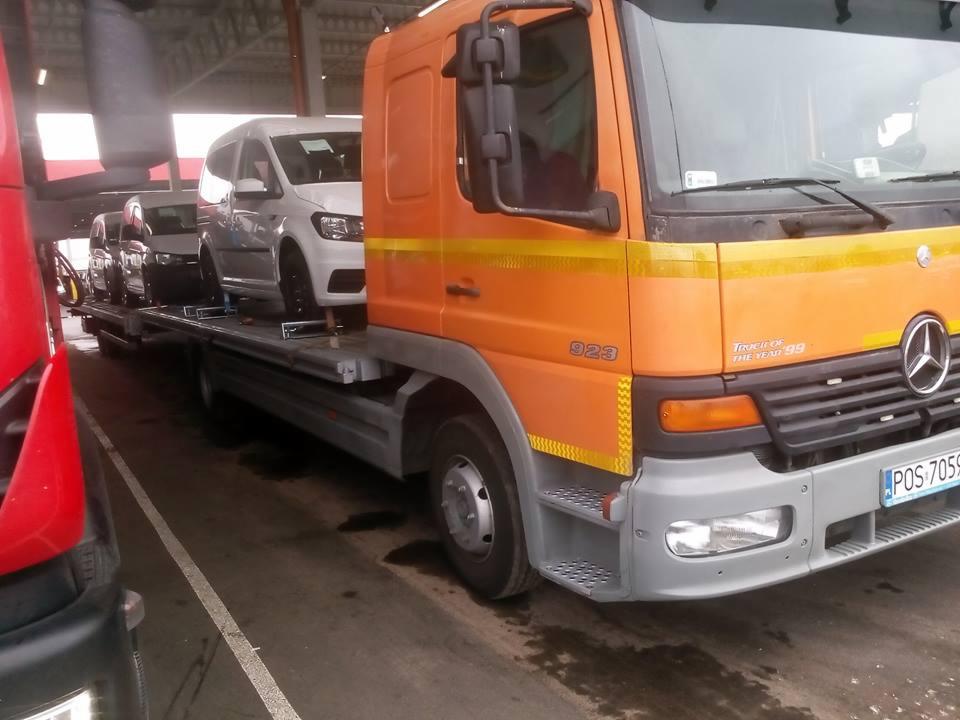 Pomarańczowa laweta Mercedes przewozi trzy samochody dostawcze, w tym białego Volkswagena Caddy, widoczny numer rejestracyjny lawety POS 7059.
