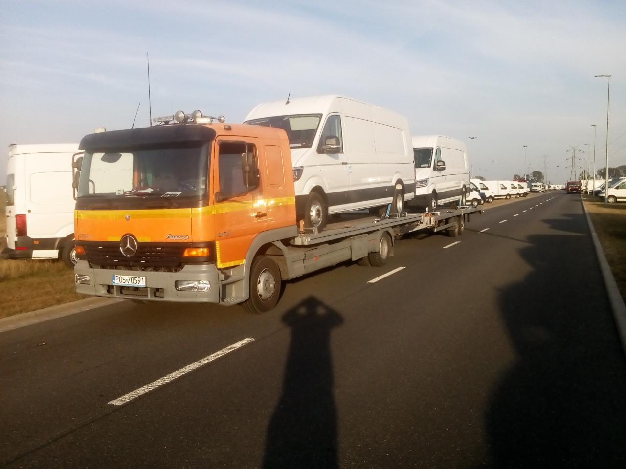Pomarańczowa laweta Mercedes-Benz Atego przewozi trzy białe busy na autostradzie w słoneczny dzień, cień fotografa widoczny na asfalcie.