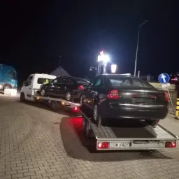 Dwa ciemne samochody osobowe na lawetach, jeden za drugim, transportowane nocą; widoczny fragment niebieskiej ciężar&oacute;wki z lewej strony i budynek w tle.