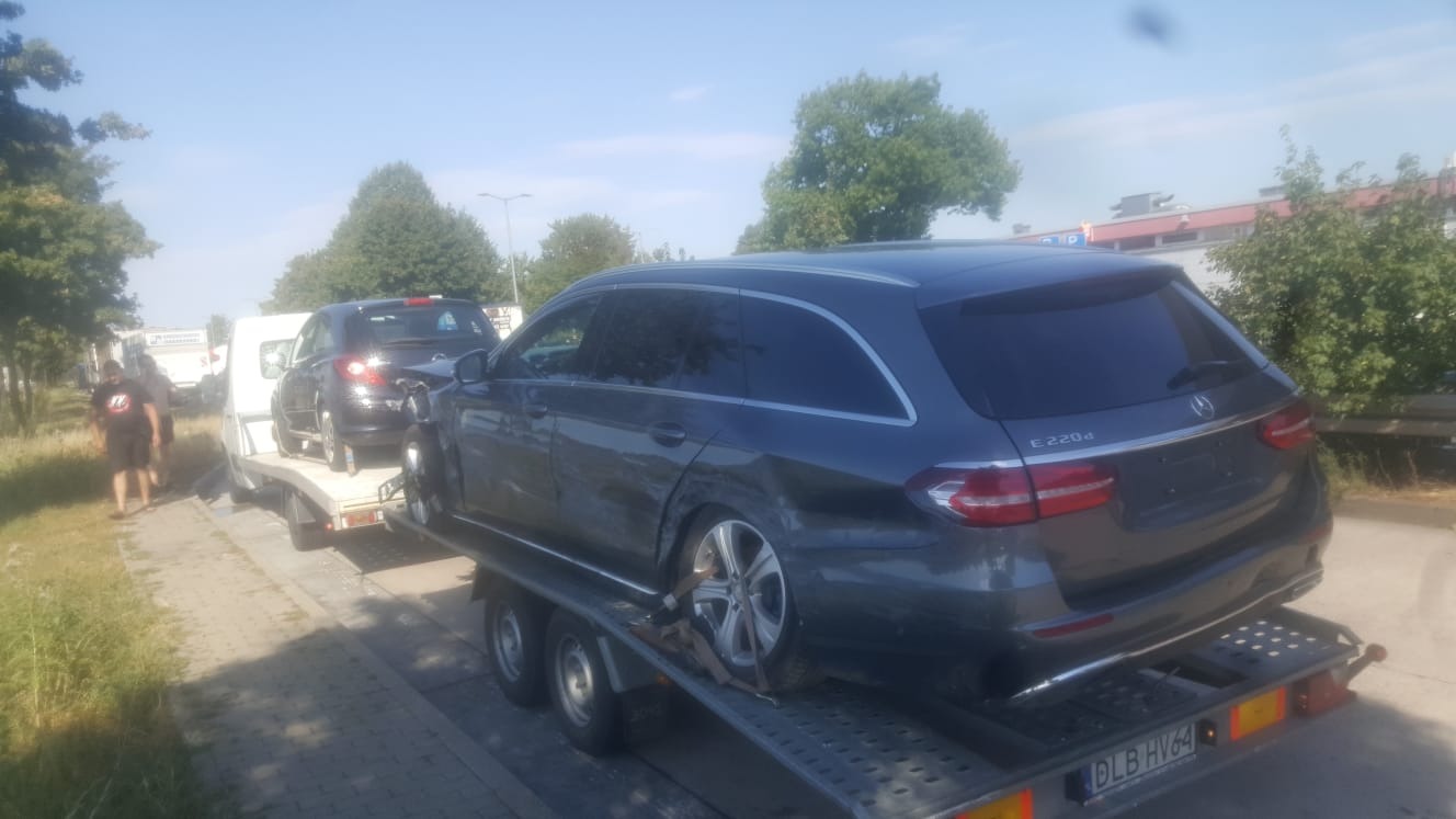 Uszkodzony, szary Mercedes E220d kombi na platformie lawety, widoczny z tyłu i boku, zarysowania na karoserii, tablica rejestracyjna DLB HV64.