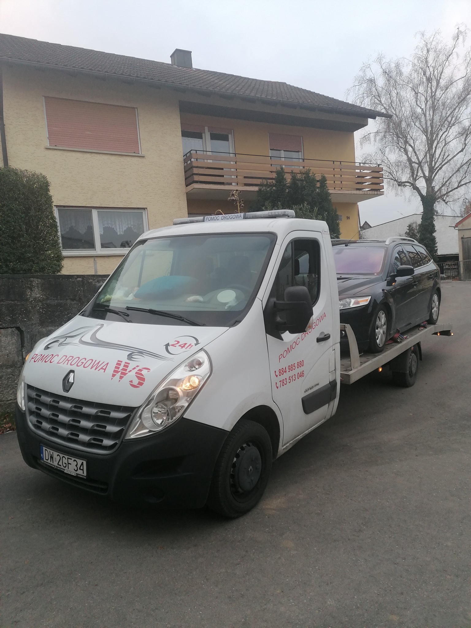 Biała laweta Renault z czarnym samochodem osobowym na platformie, zaparkowana przed budynkiem mieszkalnym z balkonem, widoczne numery telefonu kontaktowego.