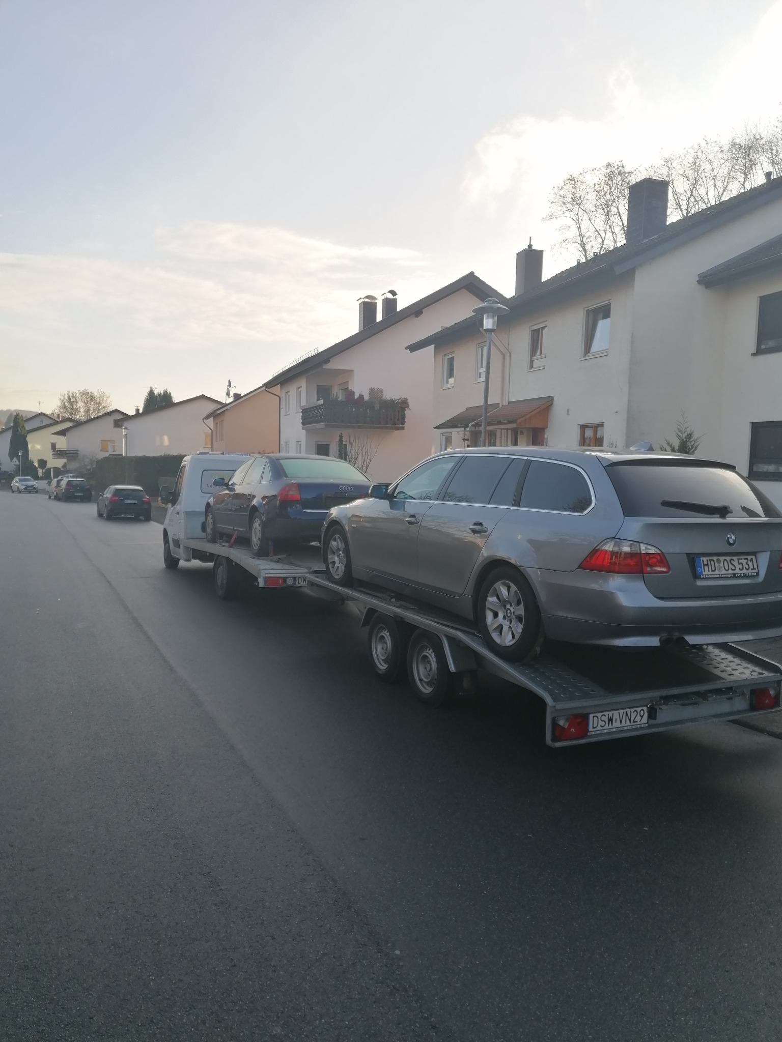 Laweta z dwoma samochodami osobowymi na platformie, zaparkowana na ulicy w dzielnicy mieszkalnej, widok z boku na szary samochód typu kombi z niemiecką tablicą rejestracyjną.