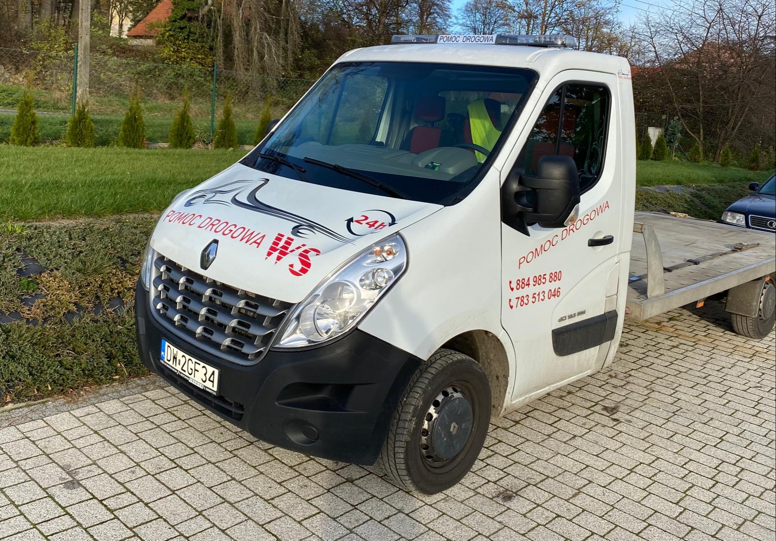 Biała laweta Renault Master z napisem 'Pomoc Drogowa' i numerem telefonu, zaparkowana na kostce brukowej w słoneczny dzień.